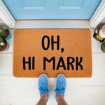 Funny welcome doormat with the text Oh Hi Mark printed in black on a natural coir fiber mat, placed on a white porch in front of a blue door with blue sneakers standing on the rug for a humorous entryway decor and housewarming
