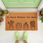 Cactus welcome doormat with funny pun text It Succs Without You Here, featuring black line art of potted succulents on a natural coir fiber mat placed on a porch, perfect for a housewarming entry or a plant lover home decor gift.