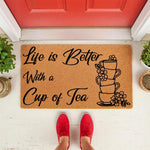 Tea lover doormat with Life is Better With a Cup of Tea text and a stack of teacups illustration, this natural coir welcome mat is a stylish front porch decor piece for tea drinkers and home entryways, shown on a white floor.