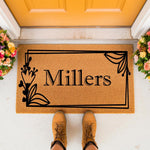 Personalized family name coir doormat featuring a black floral border design, displayed on a front porch with flowers and boots, making this custom welcome mat a stylish housewarming decor piece for any home entrance or front