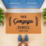 Personalized family name coir doormat featuring a custom last name and established date, displayed as a stylish outdoor welcome mat on a front porch with a blue door and green plants, perfect for housewarming or home decor.