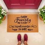 Every Family Has A Story Welcome To Ours doormat, a natural coir fiber welcome mat for front porch decor featuring black script text, placed on a concrete entryway with potted white daisy plants and a person wearing maroon