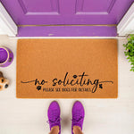 No soliciting doormat with funny dog theme, featuring black text and paw prints on a natural coir welcome mat, placed on a porch in front of a purple door to warn visitors about pets and keep solicitors away from your home