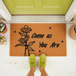 Come As You Are floral doormat featuring a black ink flower design on natural coir fiber, perfect as a front porch welcome mat for your home entrance, shown with lime green sneakers on a white floor by a green door for a stylish