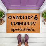 Grandkids and Grandpups Spoiled Here doormat, a natural coir fiber welcome mat for grandparents placed on a white porch, featuring bold black text that makes a fun entryway rug for any home with grandchildren and beloved dogs.