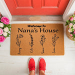 Personalized Nana welcome doormat featuring custom floral stems and family names on a natural coir fiber mat, displayed on a front porch with red shoes and flowers, making a charming housewarming decor piece for a grandmother's