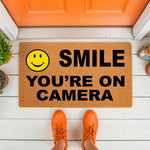 Smile You Are On Camera doormat featuring a yellow smiley face icon on natural coir fiber, perfect as a funny front porch welcome mat or outdoor entryway rug for home security, shown on a white wood porch with bright orange shoes.