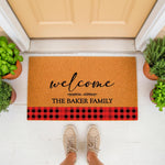 Personalized family name welcome doormat with a red and black buffalo plaid border, featuring a natural coir texture and script text, perfect for a farmhouse porch entrance or as a custom housewarming gift for a new home.