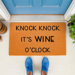Funny wine doormat with Knock Knock It's Wine O'Clock text on a natural coir fiber welcome mat, placed on a porch in front of a blue door, perfect for a home entryway or a housewarming gift for wine lovers who enjoy humorous