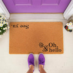 Natural coir fiber welcome doormat featuring Oh hello and See ya text with a floral design, placed on a porch in front of a purple door with person wearing purple sneakers standing on the mat for a stylish front porch decor look.