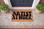 Artist at Work coir doormat featuring bold black retro typography on a natural fiber welcome mat, placed on a gray brick porch entryway with potted plants, perfect for a creative home studio or front door decor for an artist.