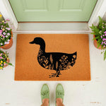 Floral duck doormat featuring a black silhouette filled with wildflowers on a natural coir welcome mat, placed at a front door entrance with potted plants and green shoes visible in this outdoor home decor entryway scene.