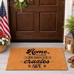 Funny Home Is Where My Bunch Of Crazies Are doormat featuring black text on a natural coir fiber welcome mat, displayed on a front porch with a dark wood door, American flag, and lantern for a stylish and humorous home entryway