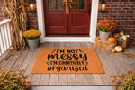 Funny quote doormat for artists featuring the text I am not messy I am creatively organized on a natural coir welcome mat, displayed on a rustic porch with fall decor including pumpkins, mums, and dried corn stalks for autumn