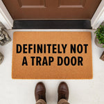 Funny doormat with the text Definitely Not A Trap Door printed in black on a natural coir fiber welcome mat, placed on a porch floor in front of a brown door for a humorous home entrance decor look that guests will surely notice.