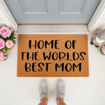 Home of the Worlds Best Mom doormat featuring natural coir fiber, placed on a front porch with pink flowers and a gnome, making a fun personalized welcome mat for a mother who loves home decor and unique entryway floor
