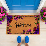 Floral Welcome Doormat with vibrant purple and pink flower designs on natural coir fiber, featuring a stylish script font on this outdoor entrance mat placed on a porch in front of a bright blue door for a colorful home entryway