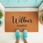 Personalized family name coir doormat featuring custom script text on a natural fiber welcome mat, placed on a front porch with teal sneakers visible, creating a stylish entryway decor piece for a new home or housewarming.