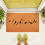 Welcome coir doormat with black script lettering and floral accents, placed on a front porch entryway.
