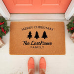 Personalized Merry Christmas doormat with pine tree icons and custom family name text, featuring a natural coir fiber welcome mat design for holiday porch decor, placed on a front porch with festive greenery and orange shoes
