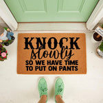 Funny Knock Slowly So We Have Time To Put On Pants doormat featuring bold black text on a natural coir fiber mat, placed on a front porch entryway as a humorous home decor welcome mat for guests and visitors to your house.