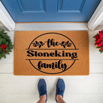 Personalized family name doormat featuring a festive holly design on natural coir fiber, perfect as a custom holiday welcome mat for your front porch entry, shown on a white floor with a blue door and seasonal greenery decor.
