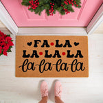 Fa La La Christmas doormat featuring black text and red dots on a natural coir welcome mat, perfect for holiday porch decor in front of a pink door with a festive wreath and poinsettia plant for a seasonal entryway floor rug look.