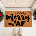 Funny Christmas doormat with black text reading merry AF, featuring holly and branch accents on a natural coir fiber welcome mat, placed on a front porch with festive greenery for a humorous holiday home decor entryway statement.