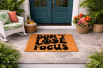 Don't Lose Focus doormat with a retro smiley face design, featuring bold black typography on a natural fiber welcome mat placed on a stone porch, perfect for a stylish front door entrance or a fun housewarming home decor gift.