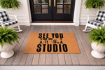 Photography studio welcome doormat featuring a camera and tripod design on natural coir fiber, perfect for a front porch entrance mat or photographer home decor, displayed on a gray wood deck with black rocking chairs and green