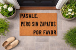 Spanish language coir doormat with black text reading Pasale, pero sin zapatos, por favor, placed on a gray tile porch with potted flowers and shoes nearby, making this a funny welcome mat for a home entrance or front door decor.