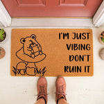 Funny frog doormat with a cute illustration of a frog drinking juice on a mushroom, featuring the text I'm just vibing don't ruin it on a natural coir welcome mat for a porch, entryway, or front door decor for frog lovers.