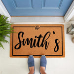 Personalized family name doormat featuring a custom last name design on natural coir fiber, perfect as a front porch welcome mat for housewarming or wedding decor, shown on a white floor in front of a blue door with a green fern