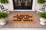 Funny Take Off Your Shoes doormat for front porch entryways, featuring bold black text on a natural coir fiber mat, perfect for a farmhouse home entrance with rocking chairs and potted ferns on a gray wood deck floor.