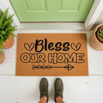 Bless Our Home doormat featuring black text and heart arrow designs on a natural coir fiber welcome mat, placed on a white porch floor in front of a green door for a stylish farmhouse entryway decor look that greets every guest.