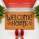Welcome Home coir doormat with black script lettering, featuring a natural fiber welcome mat for front porch decor, placed outside a red door with potted ferns, perfect for housewarming gifts and stylish entryway floor mats.