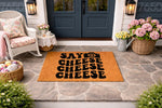 Say Cheese camera doormat for front porch entry, featuring a natural coir fiber welcome mat with bold black typography and a camera icon, displayed on a stone patio in front of a home entrance with a white wicker chair and floral