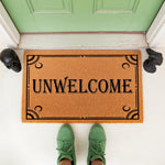 Unwelcome coir doormat with a gothic border design, featuring a funny front porch welcome mat for Halloween decor or a sarcastic home entrance, placed on a porch with a skull and raven statue for a spooky house look.