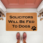 Funny dog lover doormat with the text Solicitors Will Be Fed To Dogs and a black paw print, featuring a natural coir fiber welcome mat placed on a porch floor for a humorous home entrance decor piece that dog owners will love.