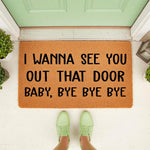 Funny welcome doormat with the text I WANNA SEE YOU OUT THAT DOOR BABY, BYE BYE BYE printed in black on a natural coir fiber mat, placed on a front porch with green shoes visible in the foreground for a humorous home entrance