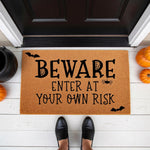 Beware Enter At Your Own Risk Halloween doormat made of natural coir fiber, featuring spooky bat and spider web designs for a festive front porch entryway decor piece that welcomes guests with a fun and scary seasonal holiday