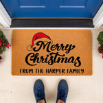 Personalized Merry Christmas doormat with a festive Santa hat design, featuring custom family name text on a natural coir fiber welcome mat for holiday porch decor, placed outside a blue front door with seasonal greenery accents.