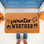 Sweater Weather fall doormat featuring black text and acorn icons on natural coir fiber, perfect for autumn porch decor, seasonal entryway rugs, and rustic farmhouse home welcome mats placed outside a blue front door with fall