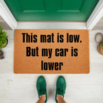 Funny car enthusiast doormat with the text This mat is low but my car is lower, featuring a natural coir fiber welcome mat placed on a porch in front of a green door, perfect for automotive fans and garage decor home entryways.
