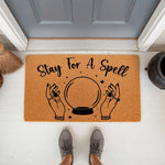 Stay For A Spell doormat featuring a crystal ball and hands design on natural coir fiber, this spooky Halloween welcome mat is a fun front porch decor piece for fall home entryways and adds a mystical touch to your outdoor floor