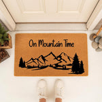 On Mountain Time doormat featuring a black mountain range and pine tree silhouette on a natural coir fiber welcome mat, perfect for outdoor porch decor, cabin entryways, or nature-inspired home entrance floor mats for hikers.
