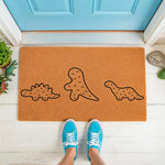 Dinosaur family doormat featuring three cute line-art prehistoric creatures on a natural coir welcome mat, placed on a porch in front of a blue door with a person wearing bright blue sneakers standing on the rug for a fun home