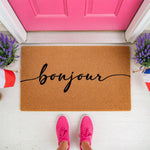 Bonjour coir doormat with black cursive script text, a natural fiber welcome mat for front porch decor, featuring a stylish French greeting design placed on a white floor in front of a bright pink door with purple lavender plants.