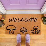 Mushroom welcome doormat with a natural coir fiber texture, featuring black line art of forest fungi and the word Welcome, perfect for a cottagecore porch decor look in front of a purple door with matching sneakers.
