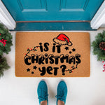 Funny Christmas doormat with Is It Christmas Yet text, a Santa hat, and holiday lights on a natural coir welcome mat, placed on a porch with pine greenery and a teal door for a festive seasonal home entrance decor look.